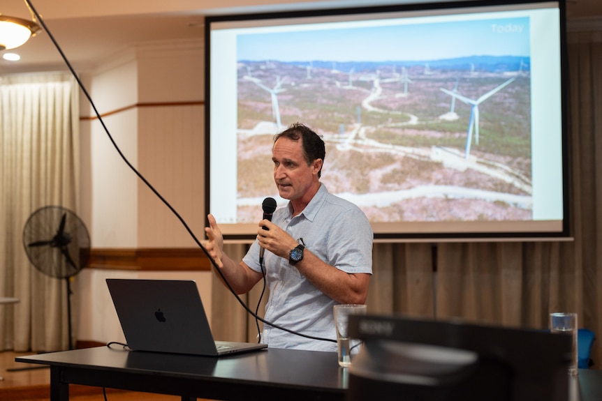 A man stands, talking into a microphone, behind him a photo of wind turbines is being projected on a screen.