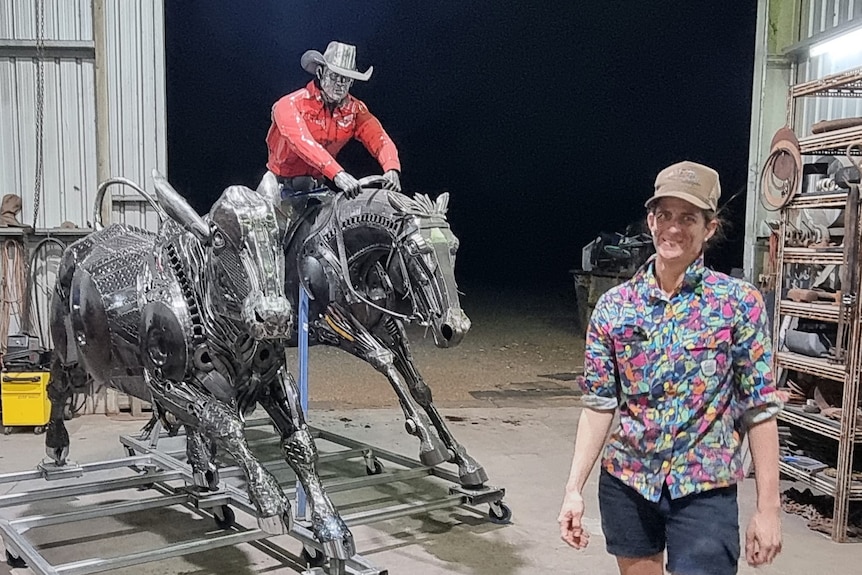 A woman in a brightly coloured shirt stands next to two sculptures of a bullock and a musterer atop a horse made from scrap meta