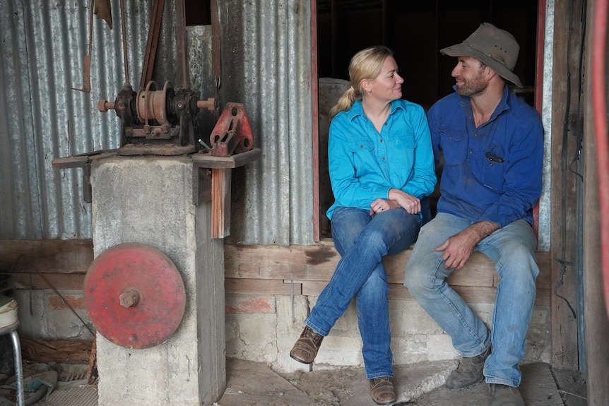 A man and a woman, both wearing long-sleeve collared shirts and jeans, look at each other seated in a shed