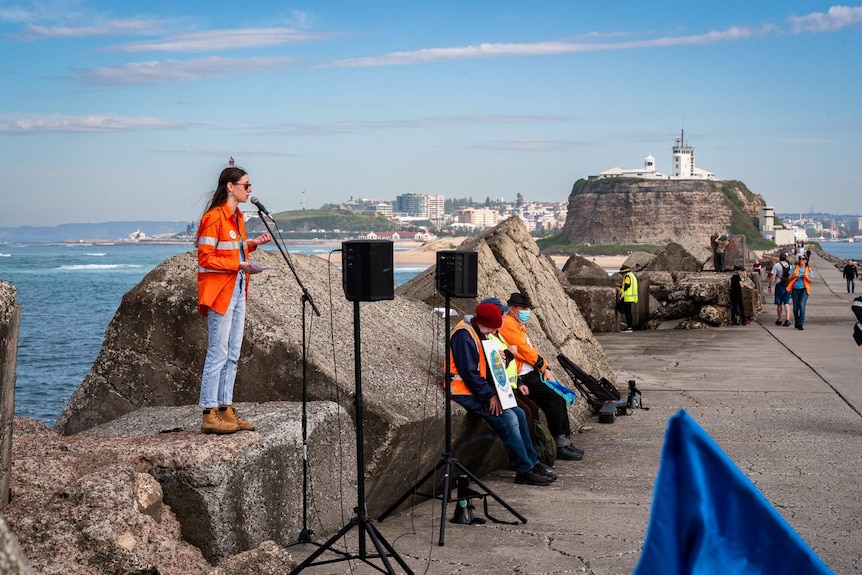 A woman with a high vis jacket on speaking into a microphone while standing on a rock that's part of a breakwall.