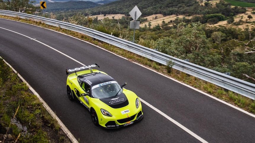 A bright yellow Lotus drives around a corner, with mountainous scenery in the background.