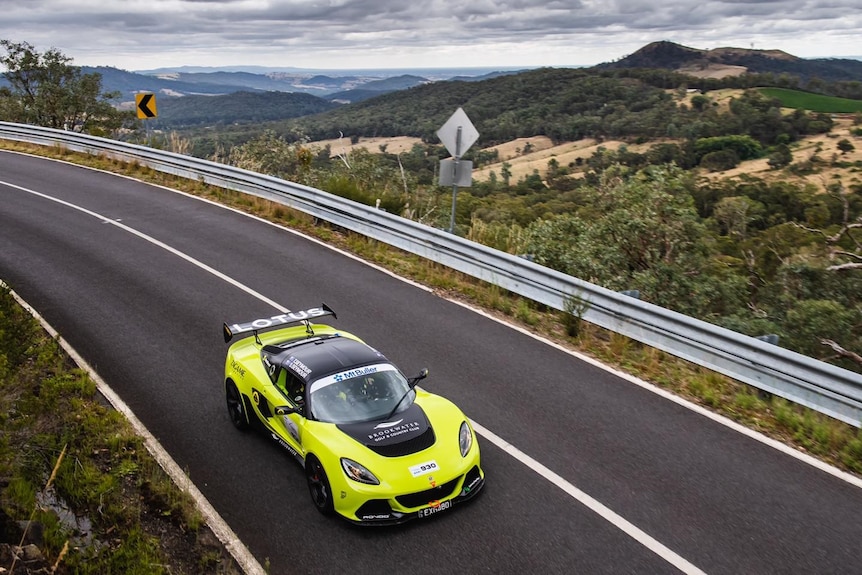 A bright yellow Lotus drives around a corner, with mountainous scenery in the background.