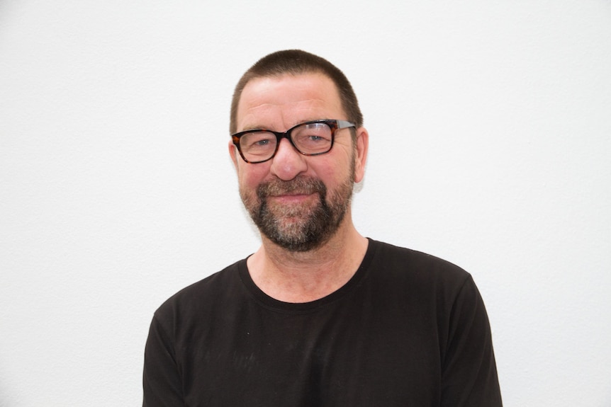 Middle-aged man with dark thick framed glasses and stubble faces camera, standing in front of a white wall.