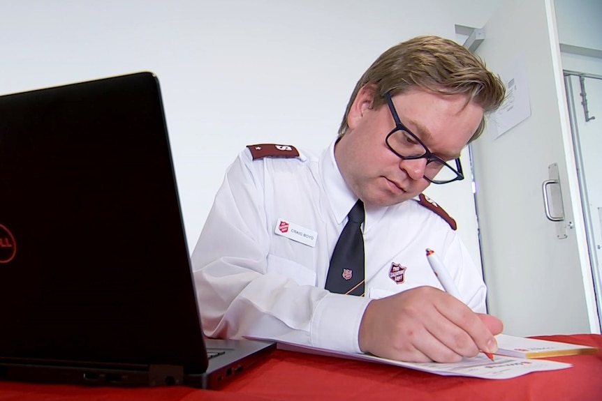 A man in Salvation Army uniform writes on a form while sitting behind a desk.