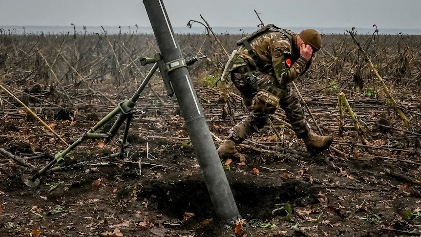 A Ukrainian soldier is pictured in a bending over with his hands over his ears as he walks away from a mortar being fired.