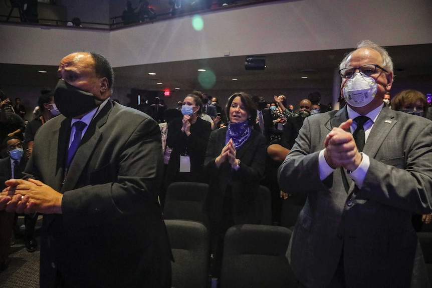 An African American man wearing a black mask and dressed in a suit stand with his hands folded next to another