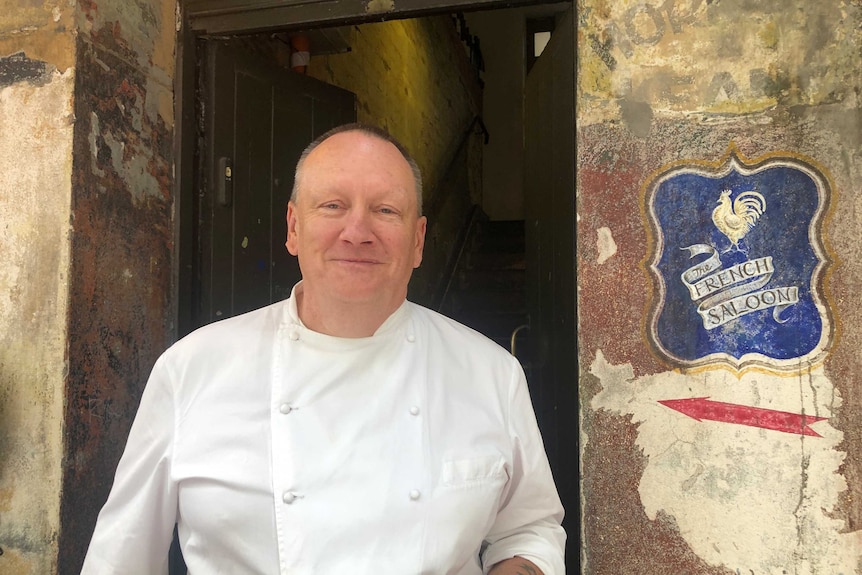 A man stands in front of an older brick building wearing a chef uniform , sign reads "The French Saloon"
