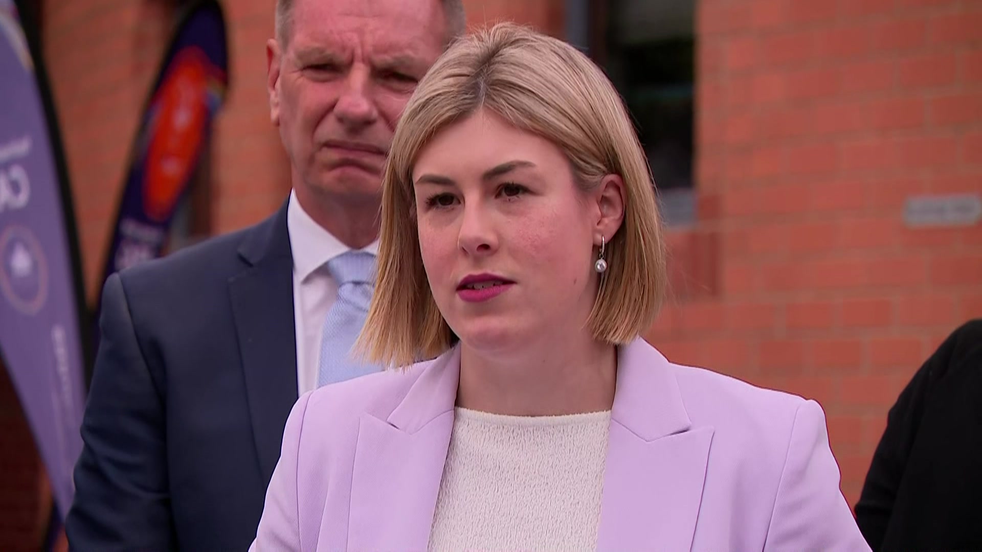 Un hombre con una chaqueta azul oscuro, camisa blanca y corbata azul claro está detrás de una mujer rubia con una chaqueta morada y un top blanco.