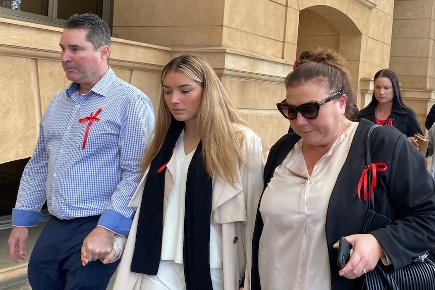 A man and two women wearing red ribbons walk, holding hands, outside a court building