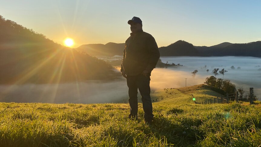 A man stand on a hill with fog in the background below and the sun creeping up behind hills in the distance.