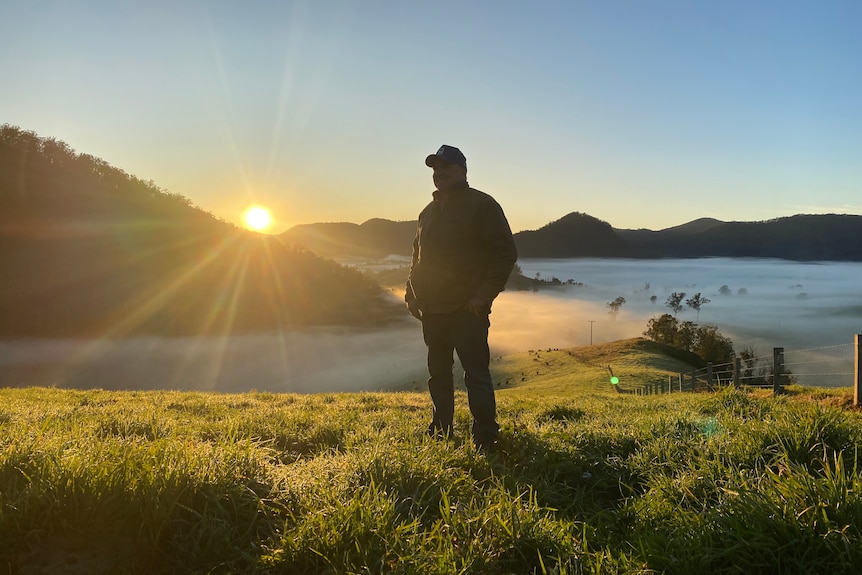 A man stand on a hill with fog in the background below and the sun creeping up behind hills in the distance.