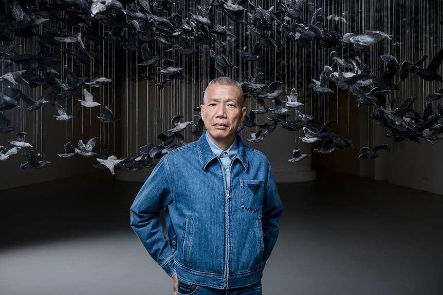 Colour photograph of artist Cai Guo-Qiang standing in a gallery space in front of an artwork featuring a flock of birds.