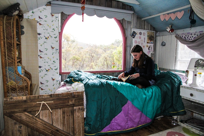 A girl in school uniform sits on her bed beside a large window, reading.