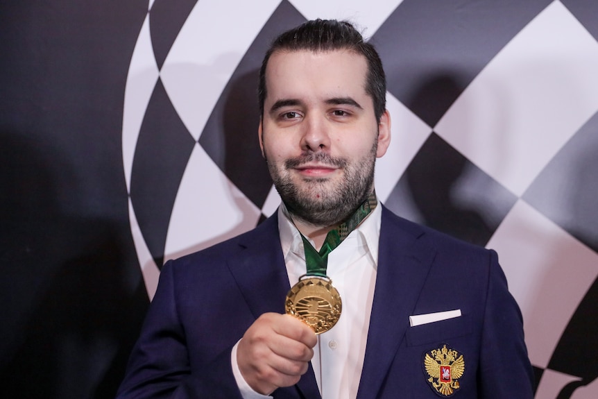 A chess Grandmaster smiles as he holds the gold medal hanging around his neck after winning a tournament.