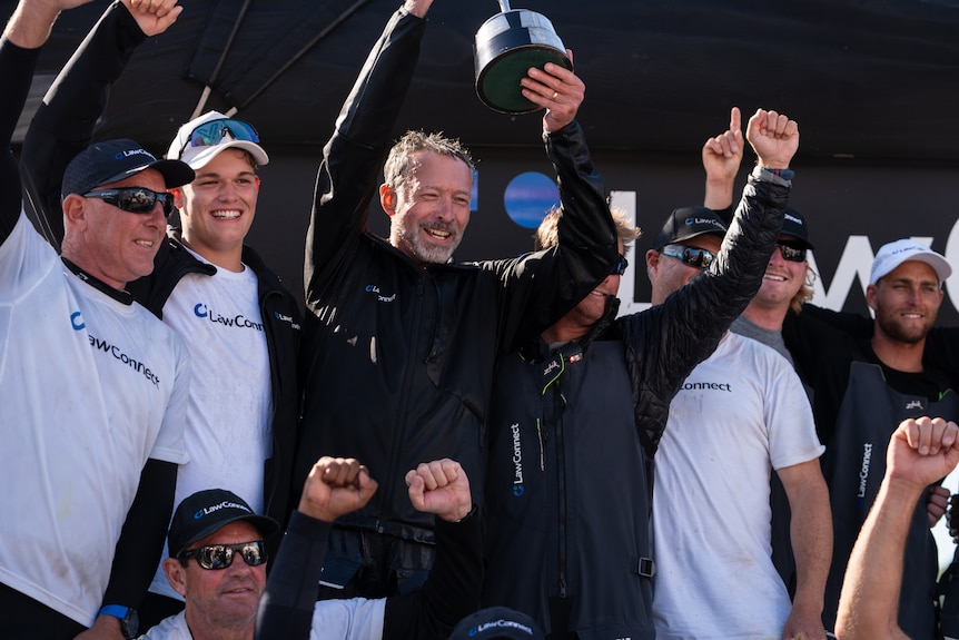 Crew of the yacht Lawconnect smile and hold up trophy, the black sails of their yacht in the background.