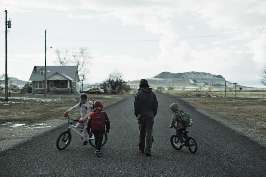A man and three children (two of which are on bikes) on a street in a desolate town, patches of snow