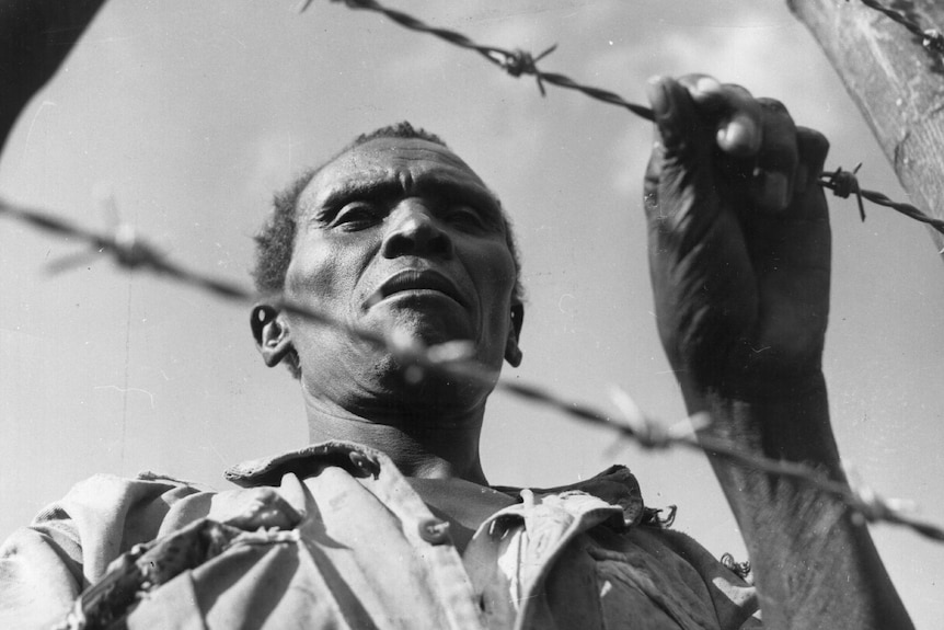 An African man with a sombre look grabs a barbed wire fence he's standing behind.