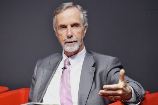 A man with a grey bear sits on a red couch, wearing a grey suit. 