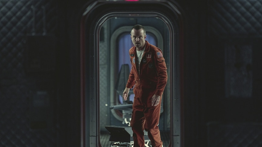 A man wearing an orange spacesuit walks through a dark tunnel