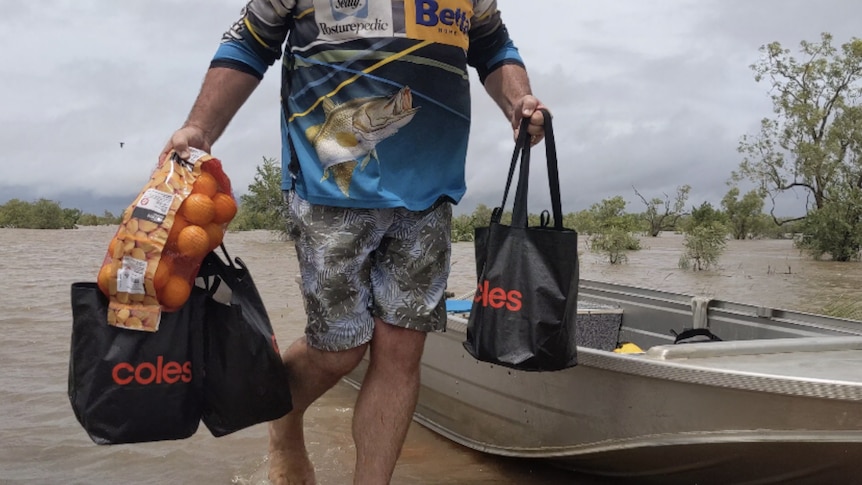 A man in a fishing shirt carries two shopping bags in front of a tinny and floodwaters