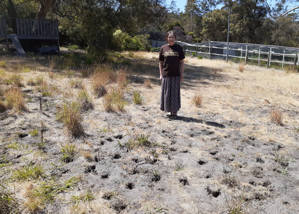 A woman stands smiling in a shirt that says worlds ugliest lawn. Her lawn has patches of grass, dirt and holes dugs by wildlife.