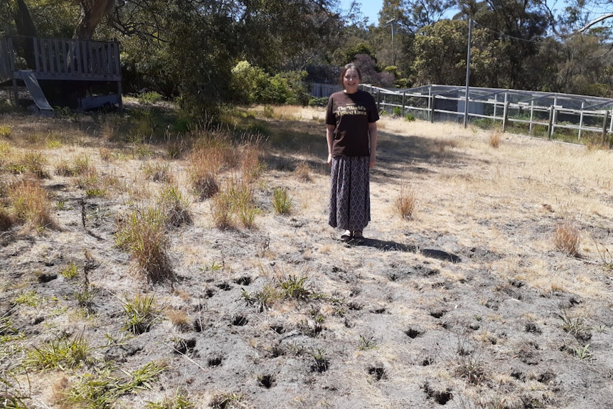 A woman stands smiling in a shirt that says worlds ugliest lawn. Her lawn has patches of grass, dirt and holes dugs by wildlife.