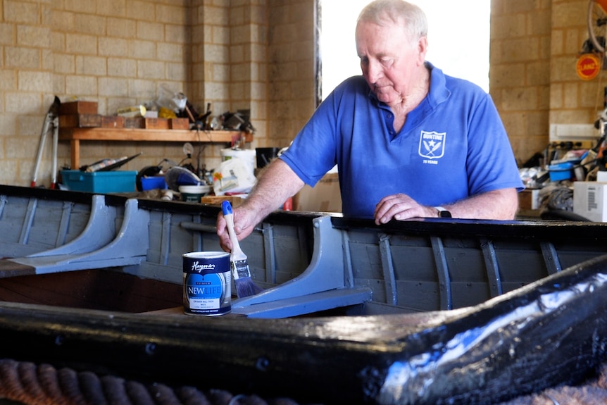 An elderly man leaning over an old boat painting it, in a shed.