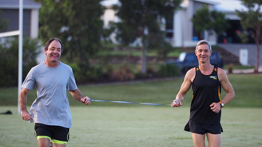 Blind runner Robin Braidwood runs alongside coach Norbert Petras on a grass field. They are joined by a tether and smiling.