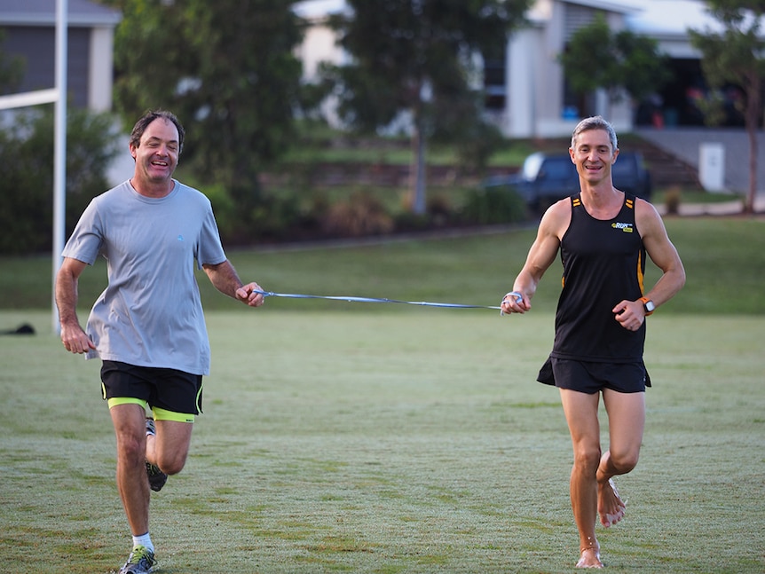Blind runner Robin Braidwood runs alongside coach Norbert Petras on a grass field. They are joined by a tether and smiling.
