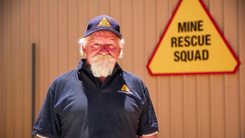 A man with a white beard stands looking serious in front of a sign that says 'Mine Rescue Squad'