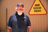 A man with a white beard stands looking serious in front of a sign that says 'Mine Rescue Squad'