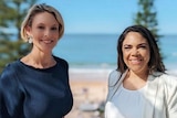 Two women smiling at the camera, with a beach and ocean behind them,