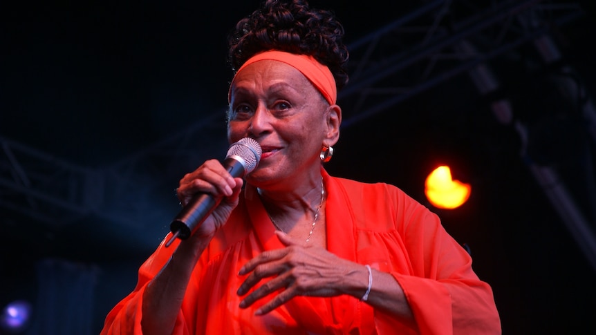 A colour photo of an older Omara Portundo in an orange dress, singing into a hand-held mic