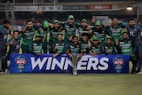 A group of Pakistan cricketers pose with a silver trophy behind a banner saying "Winners".