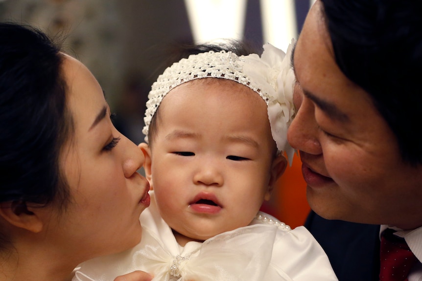 A baby wearing a white headband receives kisses on the cheek from her mother and father on each side. 