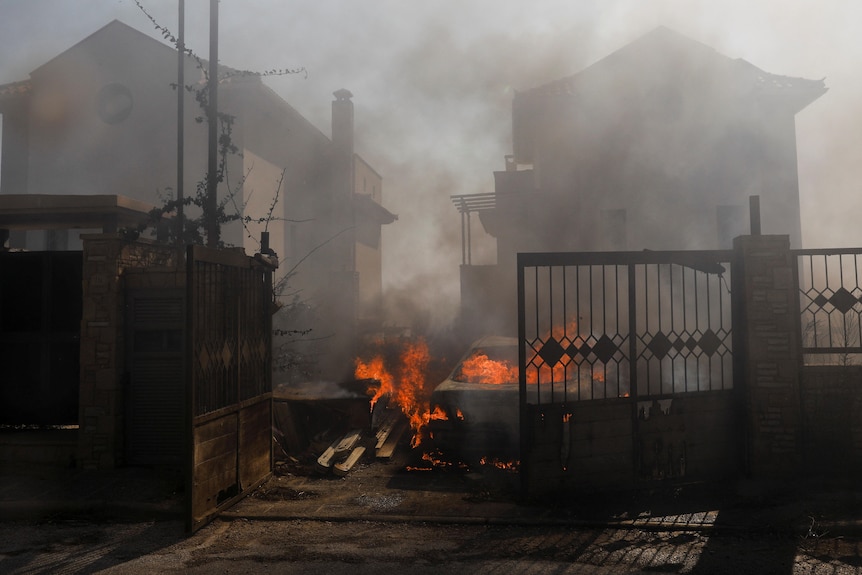 A car engulfed in flames between two houses covered in smoke. 