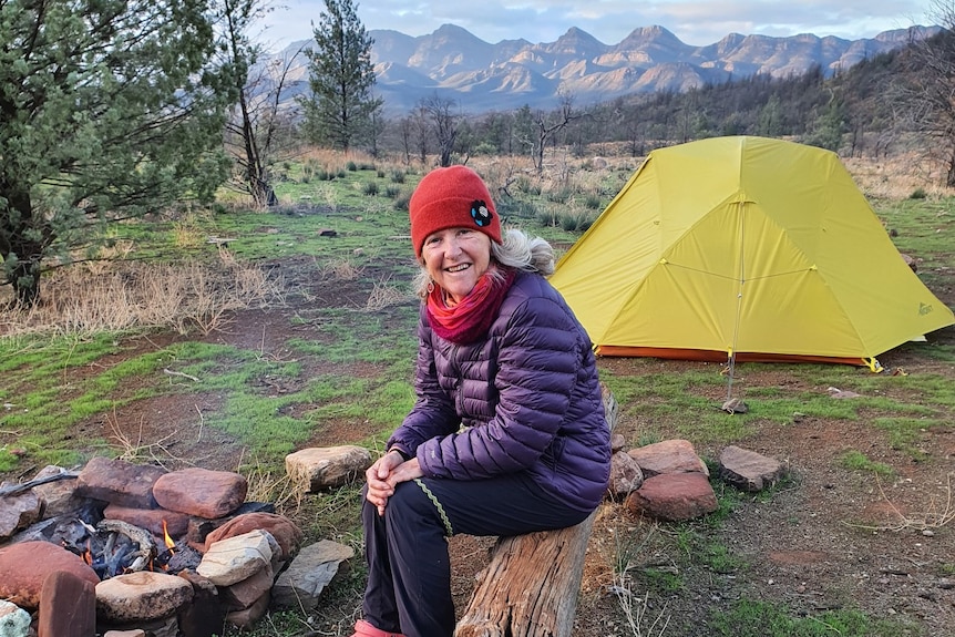 A woman sits in front of a tent, near a camp fire, wearing a beanie and warm clothes with mountains in the background