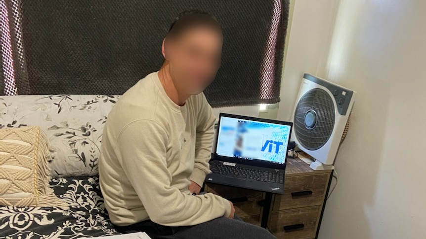 A man sits on the edge of a bed with an open laptop on the small table beside him. His face is blurred.