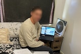 A man sits on the edge of a bed with an open laptop on the small table beside him. His face is blurred.