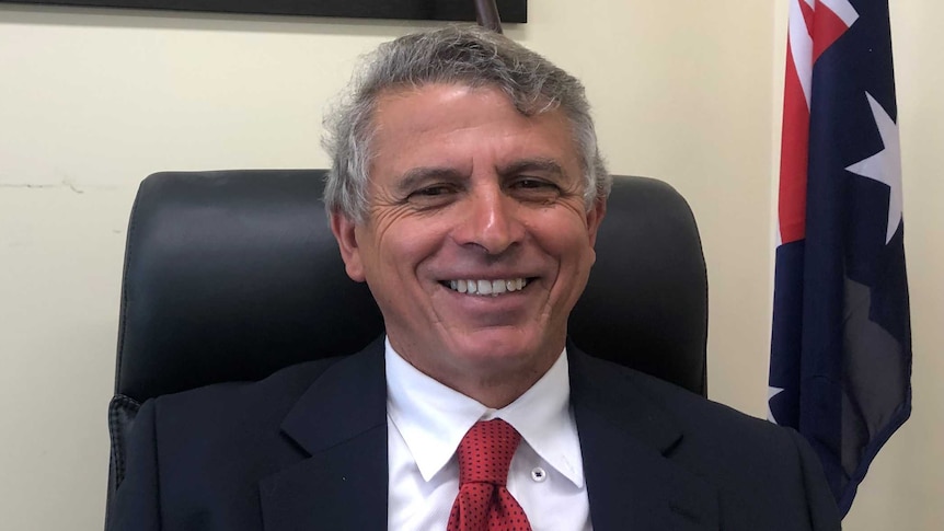 Man in suit and red tie sits in desk chair in office, smiling.