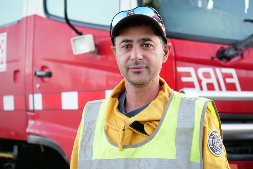 A man in a yellow fire uniform and high vis vest wearing a cap stands in front of a fire truck and looks at the camera.
