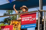 A woman in a red shirt and hat stands on a stage under an Australian flag and waves to a crowd.