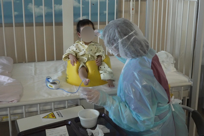 A baby sits on a cot in a feeding chair while a nurse in full PPE spoons food into her mouth 