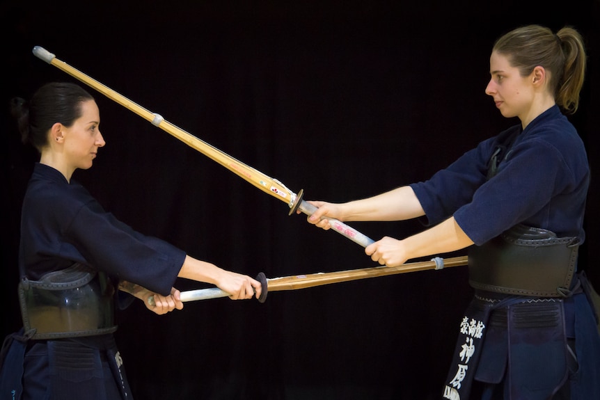 Two female kendoka without their protective helmets on stand face-to-face while holding bamboo swords.