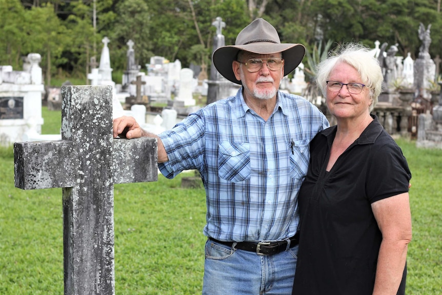 A man and woman stand next to a weathered old cross headstone in a cemetery.