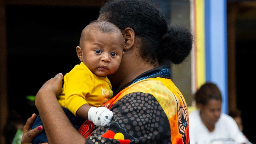A baby in a yellow t-shirt being held by a woman.