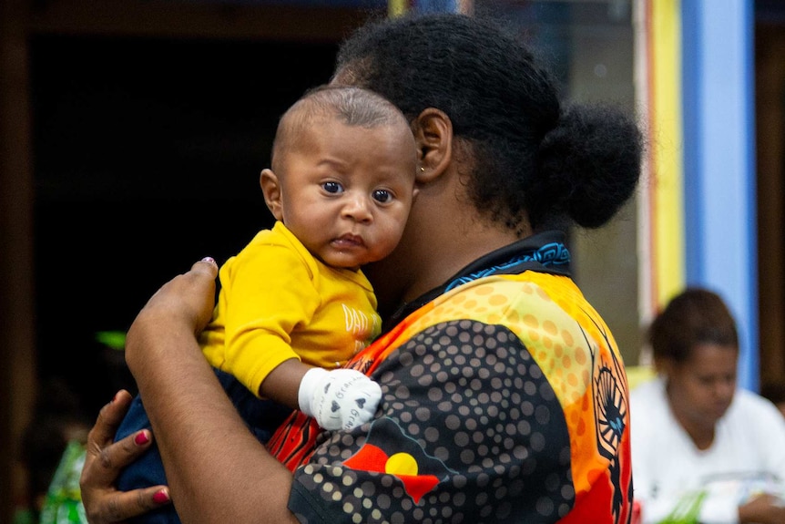 A baby in a yellow t-shirt being held by a woman.