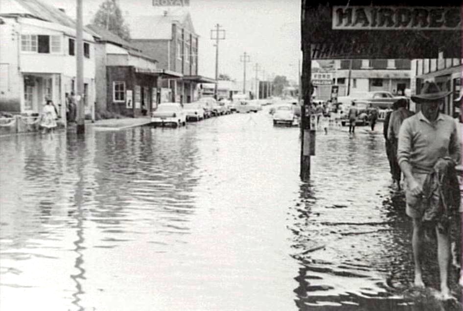 A black and white photo of people wading through water outside shops on a flooded street.