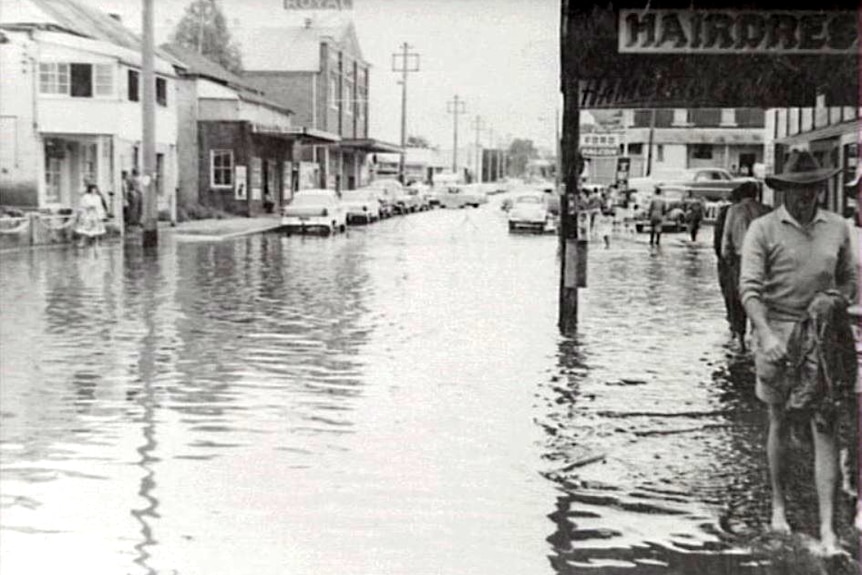 A black and white photo of people wading through water outside shops on a flooded street.