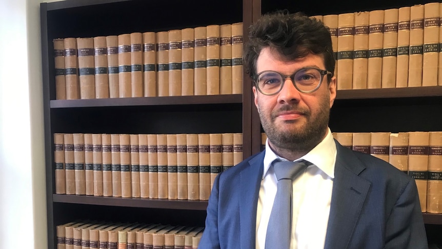 A man in a suit stands in front of a book case, he wears classes.